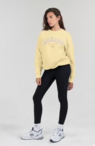 Woman wearing a yellow sweatshirt with 'Monaco' text, black leggings, and white sneakers on a gray background