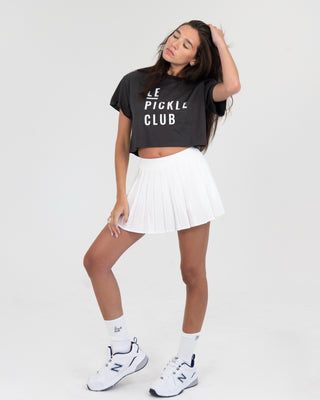 Woman wearing a black 'LE PICKLE CLUB' t-shirt, white pleated skirt, and white sneakers on a white background