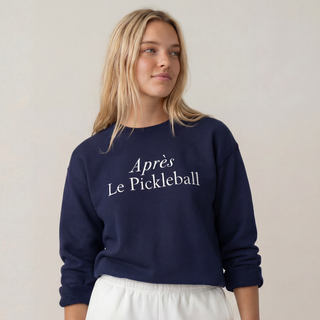 Woman wearing a navy sweatshirt with 'Apres Le Pickleball' text and white shorts on a beige background