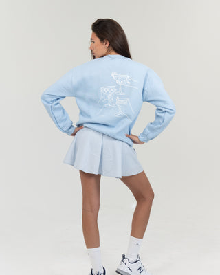 Person wearing a light blue sweatshirt  Apres le Pickleball crewneck and skirt set with a white logo on a plain background