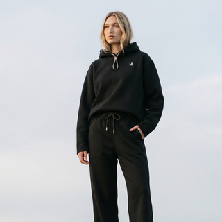 Person wearing a black hoodie and pants with a logo, standing against a light background