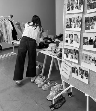 Founder, Ashley,  at a photoshoot  looking at clothing with a bulletin board displaying photos in the foreground.