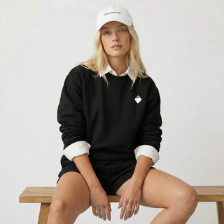 Woman wearing a black sweater with a white collar and a small logo, sitting on a wooden bench against a plain background.