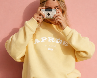 Blonde woman wearing a yellow outfit with 'Apres le pickle' printed on it, standing against a pink background.